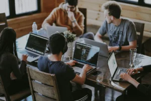 Grupo de jovens trabalhando cada um com seu notebook em volta de uma mesa de madeiras, sentados em cadeiras de madeira escura.