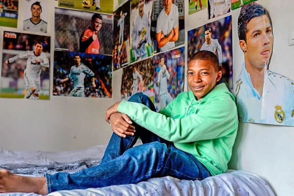Foto do Mbappé quando criança sentado sobre uma cama. Posteres do Cristiano Ronaldo na parede do seu quarto se destacam ao fundo.