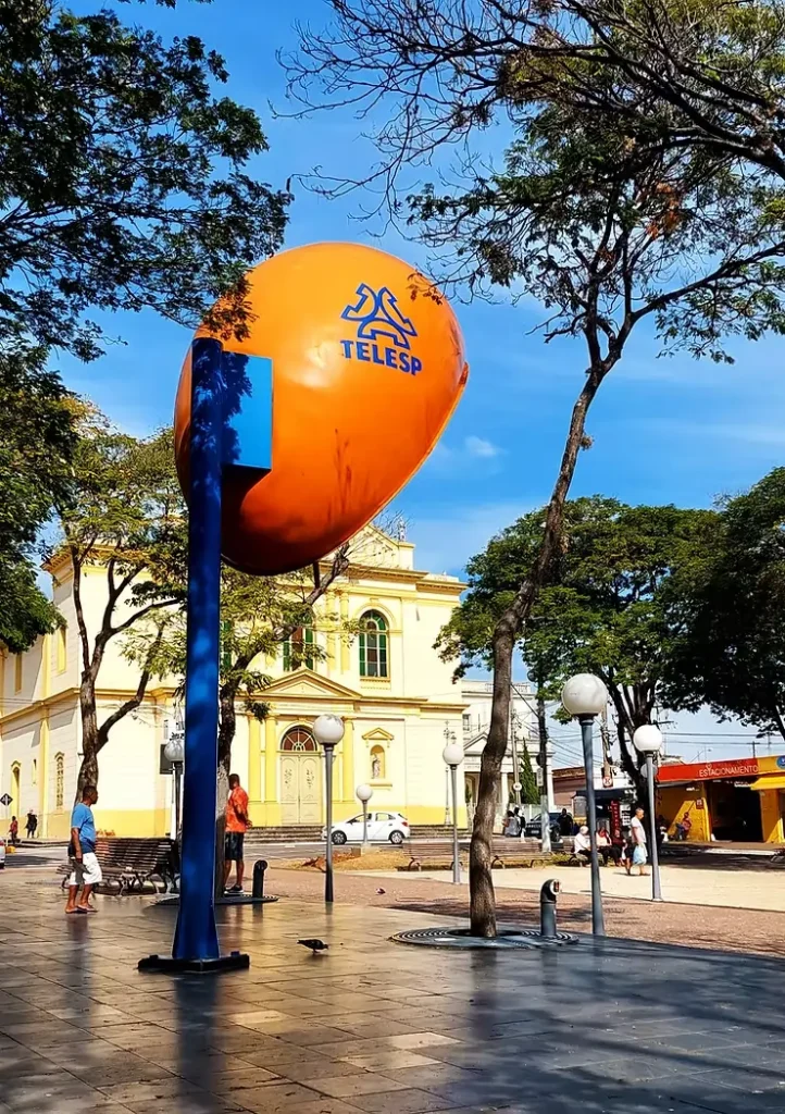 Orelhão gigante da cidade de Itú. Fixado numa praça da cidade, ao fundo se vê árvores e um edifício antigo.