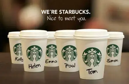 Foto de vários copos da Starbucks mostrando os nomes escritos em cada copo, como Tom e Helen. A frase de destaque no banner fala "We're Starbucks." ou em português "Nós somos Starbucks".